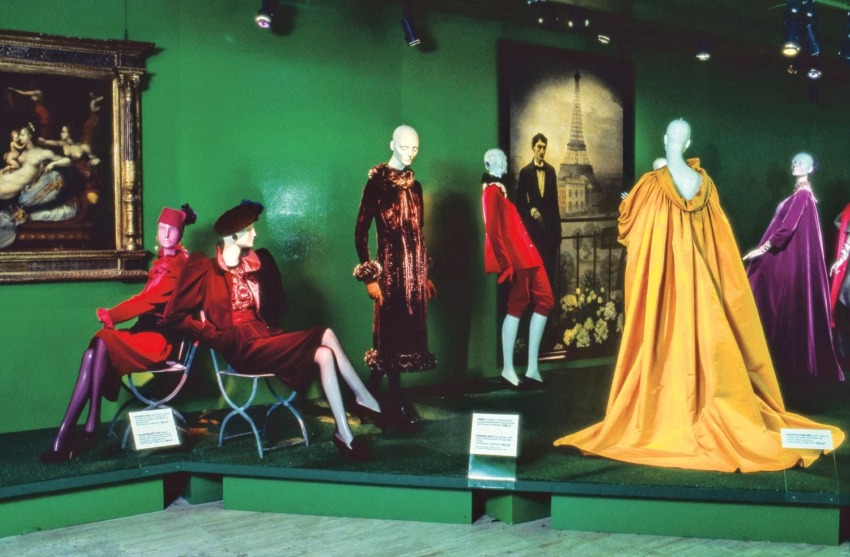 Fashion exhibit showcasing vibrant mannequins in colorful attire against a green backdrop with paintings.
