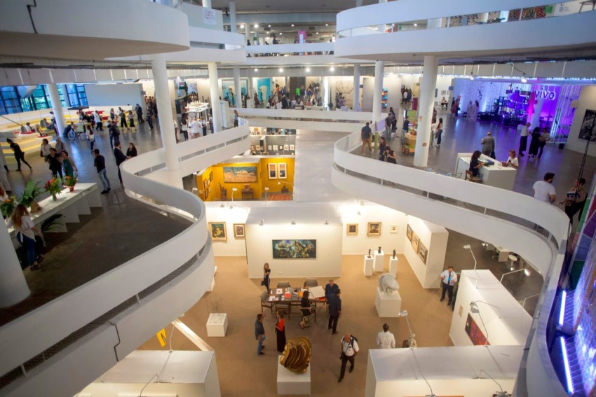 Busy modern art gallery with people exploring various exhibits on multiple levels, featuring paintings and sculptures.
