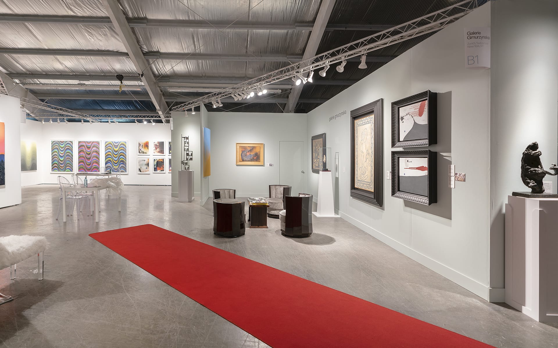 Art gallery interior with modern paintings on walls, red carpet, contemporary seating, and bright lighting.