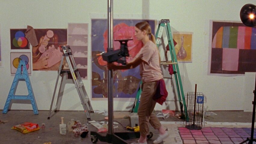 Artist in a studio adjusting equipment with colorful artwork and ladders in the background.