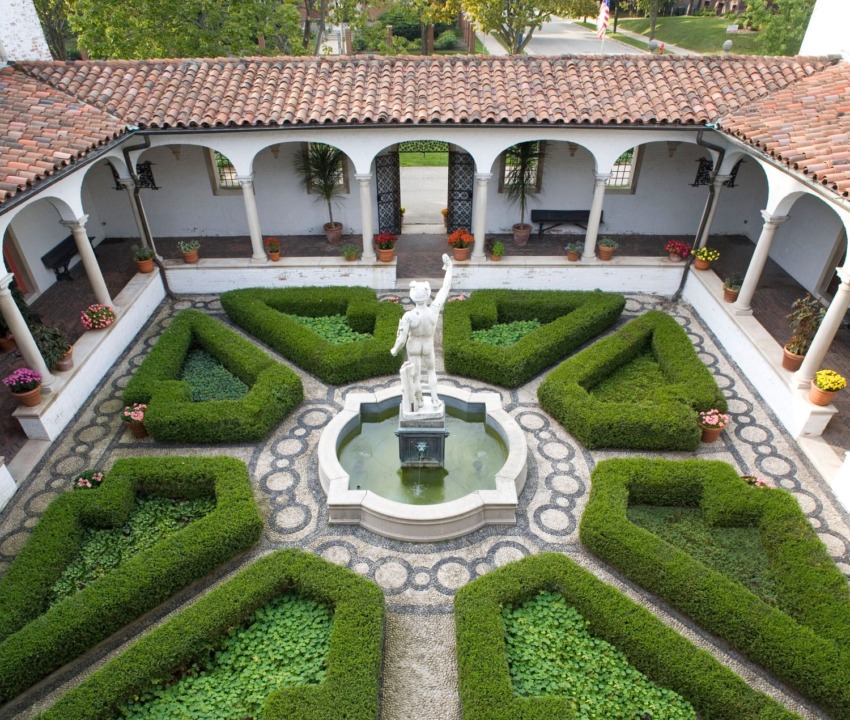 Courtyard garden with topiary, fountain, and statue surrounded by a covered walkway and colorful flowers.