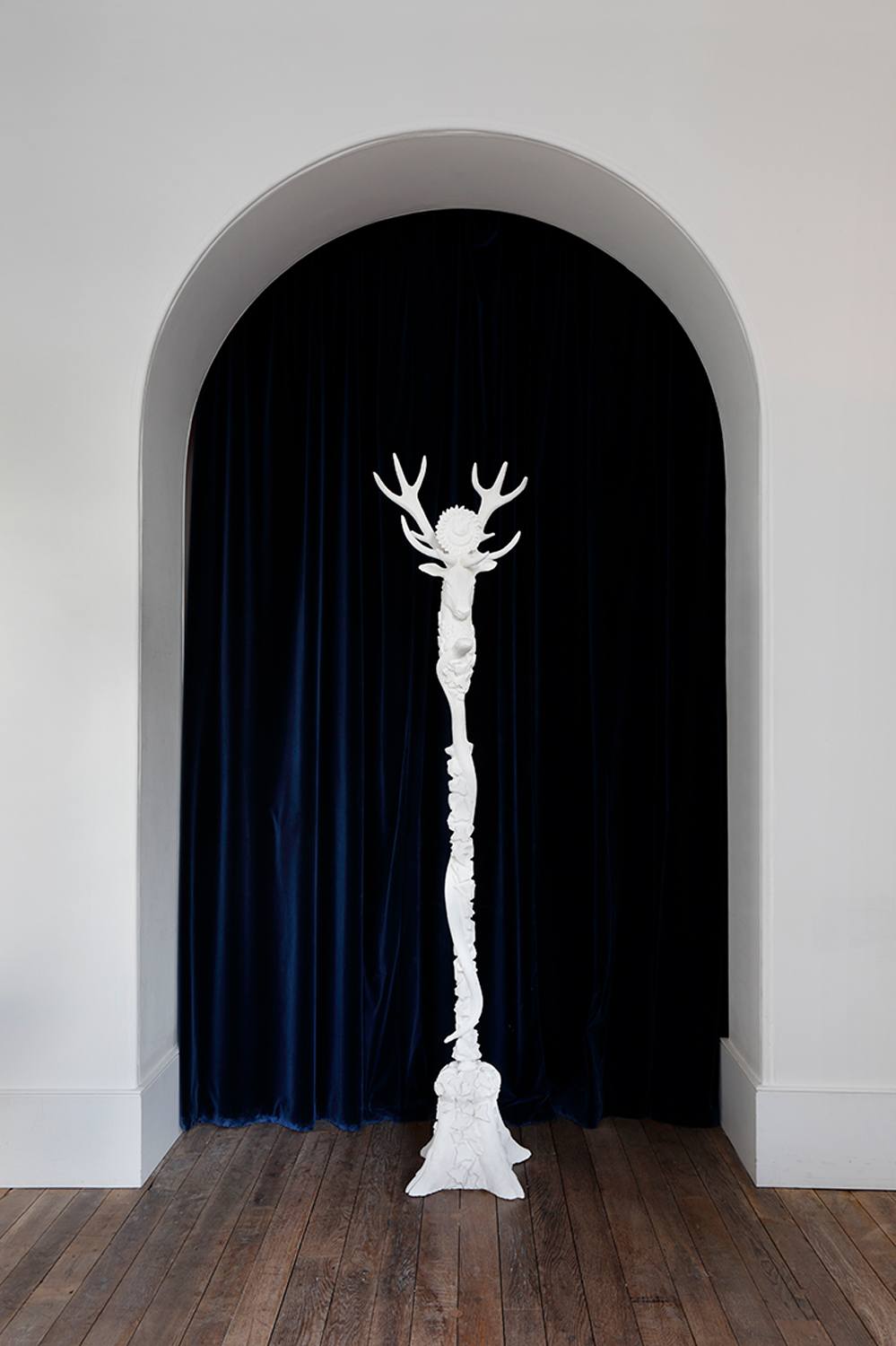 White sculptural artwork resembling an elongated figure with antlers set against dark blue curtains in an arched alcove.