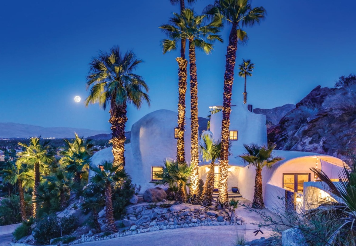 Desert home with unique architecture surrounded by tall palm trees, moon rising in the evening sky in the background.