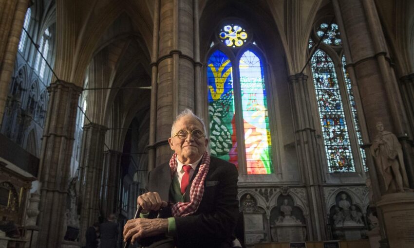 Elderly man sitting in a cathedral with colorful stained glass windows in the background.
