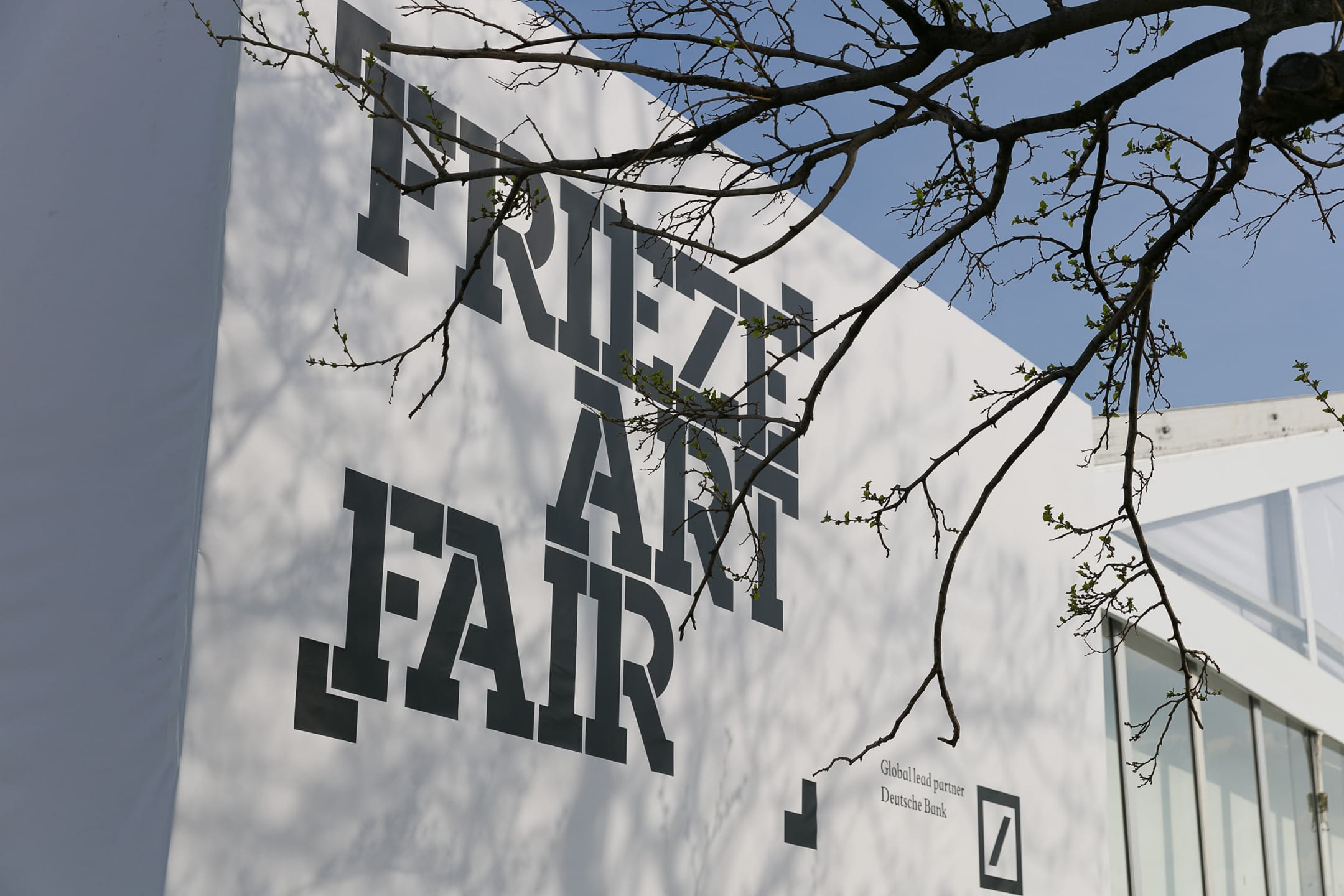 Frieze Art Fair sign on a large white structure with tree branches in the foreground and a clear sky background.