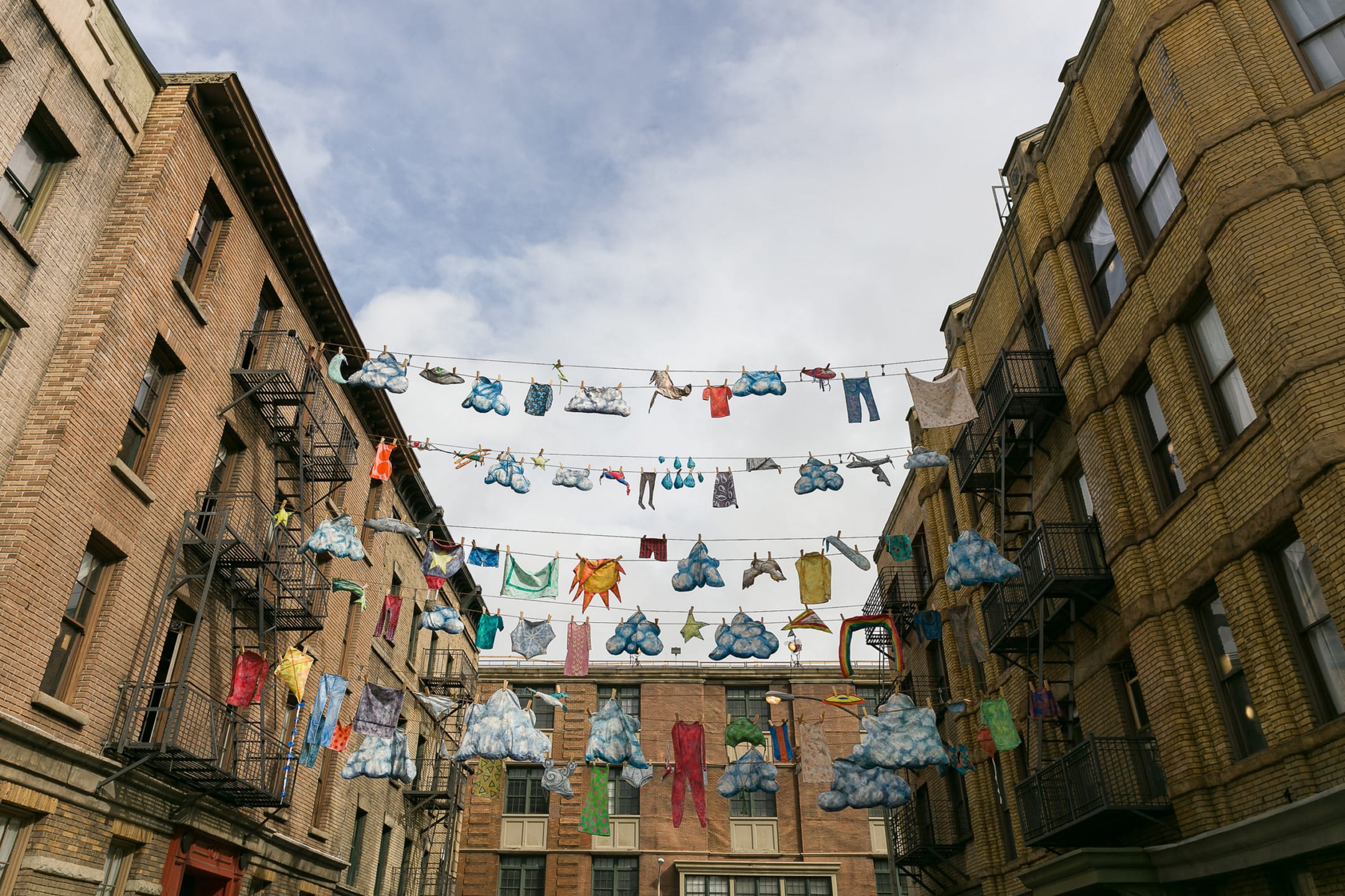 Clothes and plush items hanging on multiple clotheslines strung between brick apartment buildings under a cloudy sky.