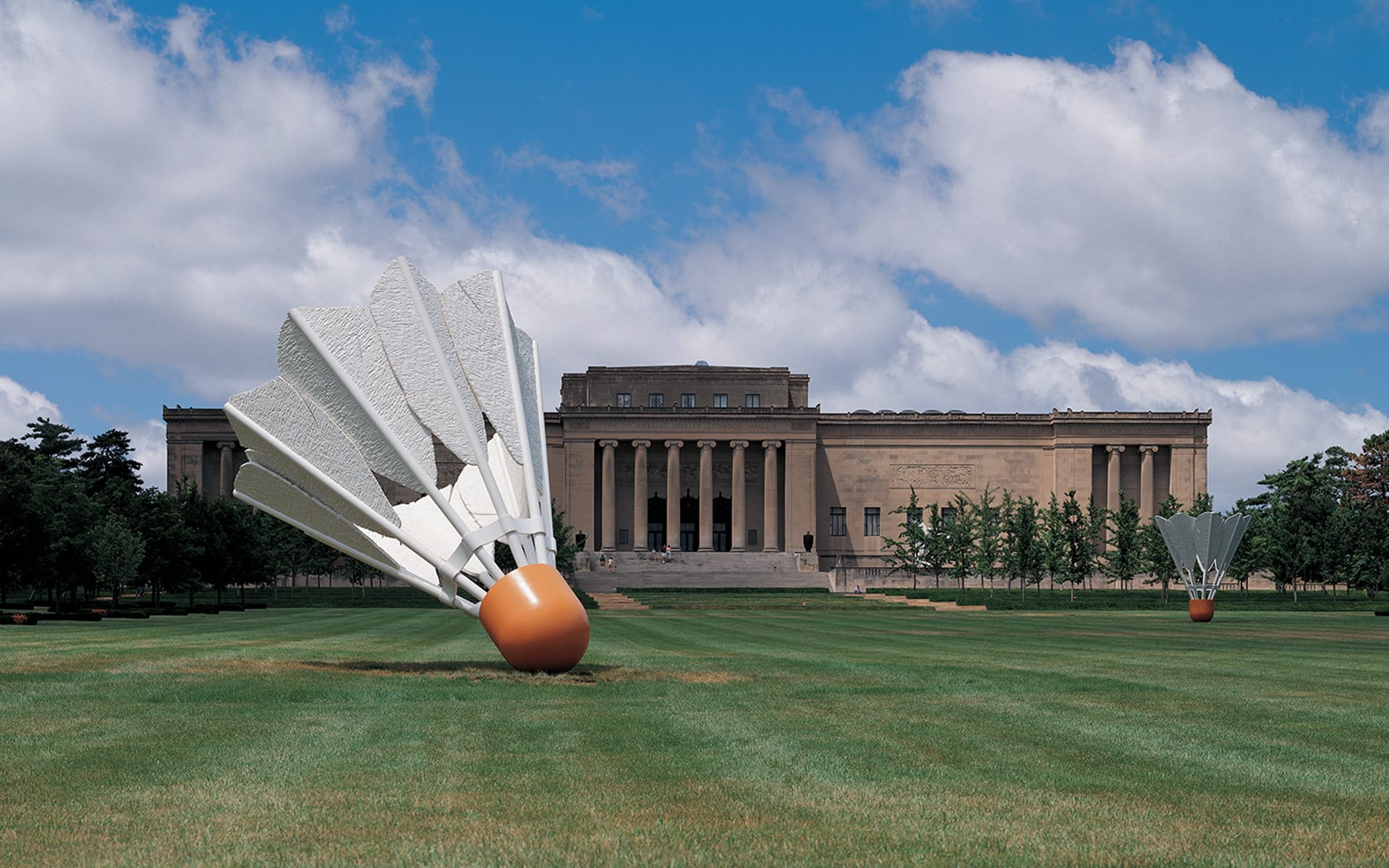 Large shuttlecock sculptures on a grassy field in front of a historic building with columns under a blue sky with clouds