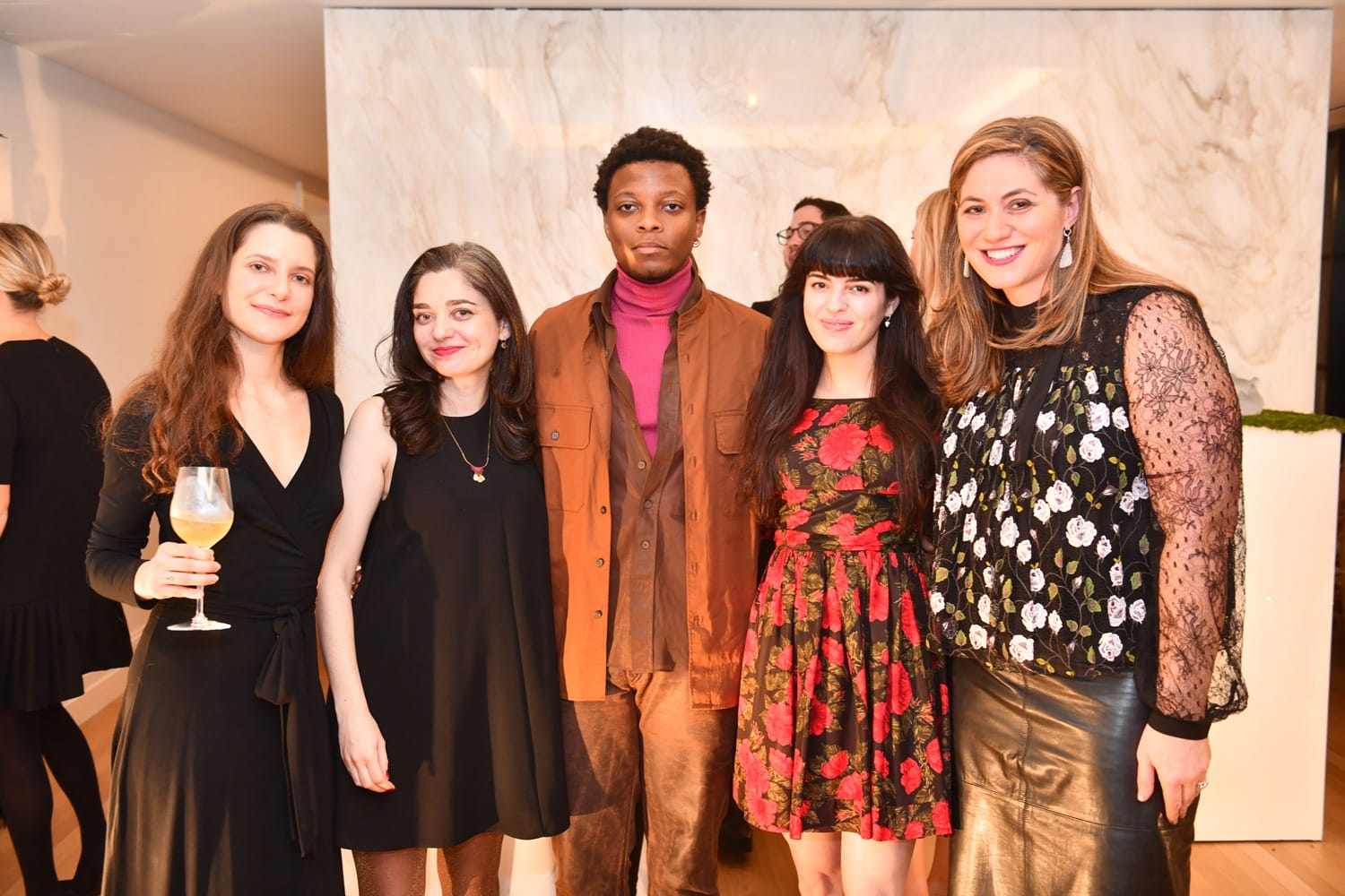 Group of five people smiling and posing at an indoor event, with three women in black dresses and two people in patterned outfits.