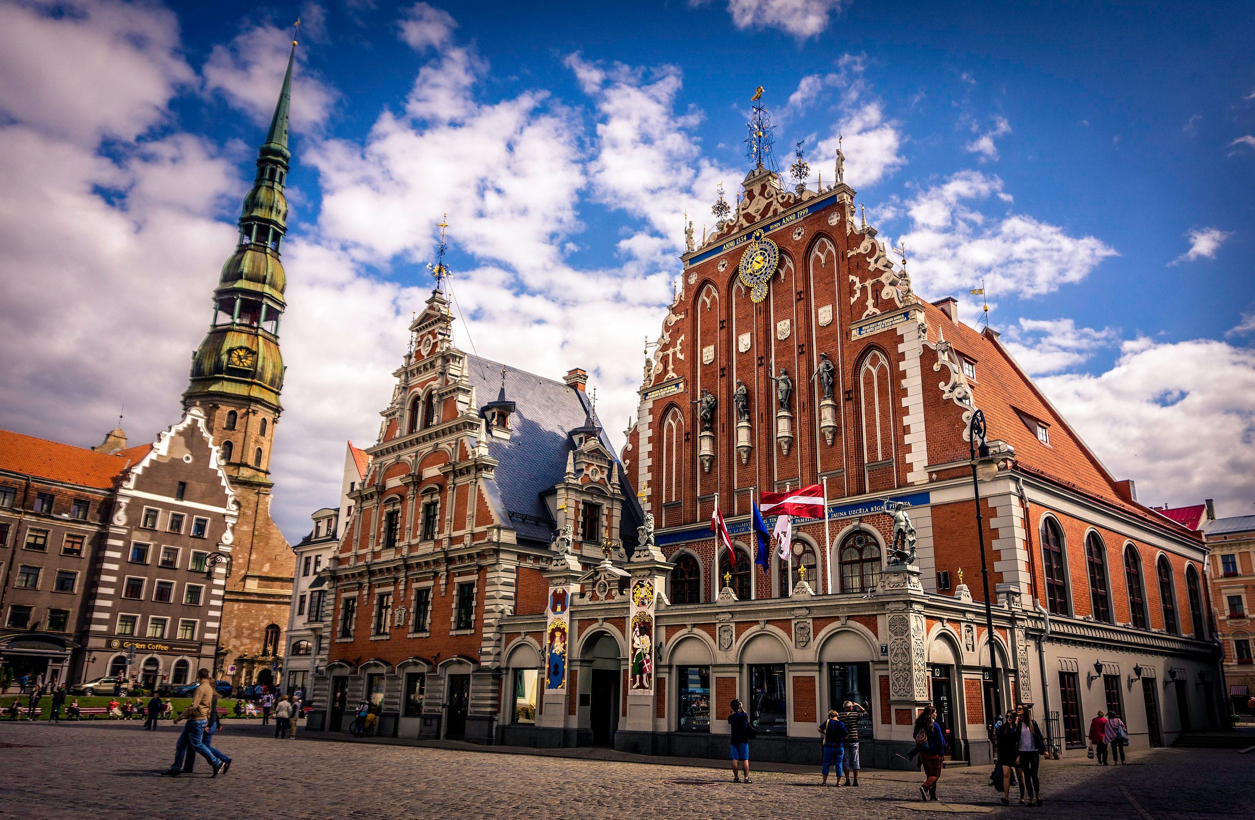 Historic red brick building with ornate details in a vibrant city square, under a blue sky with fluffy clouds, people walking.
