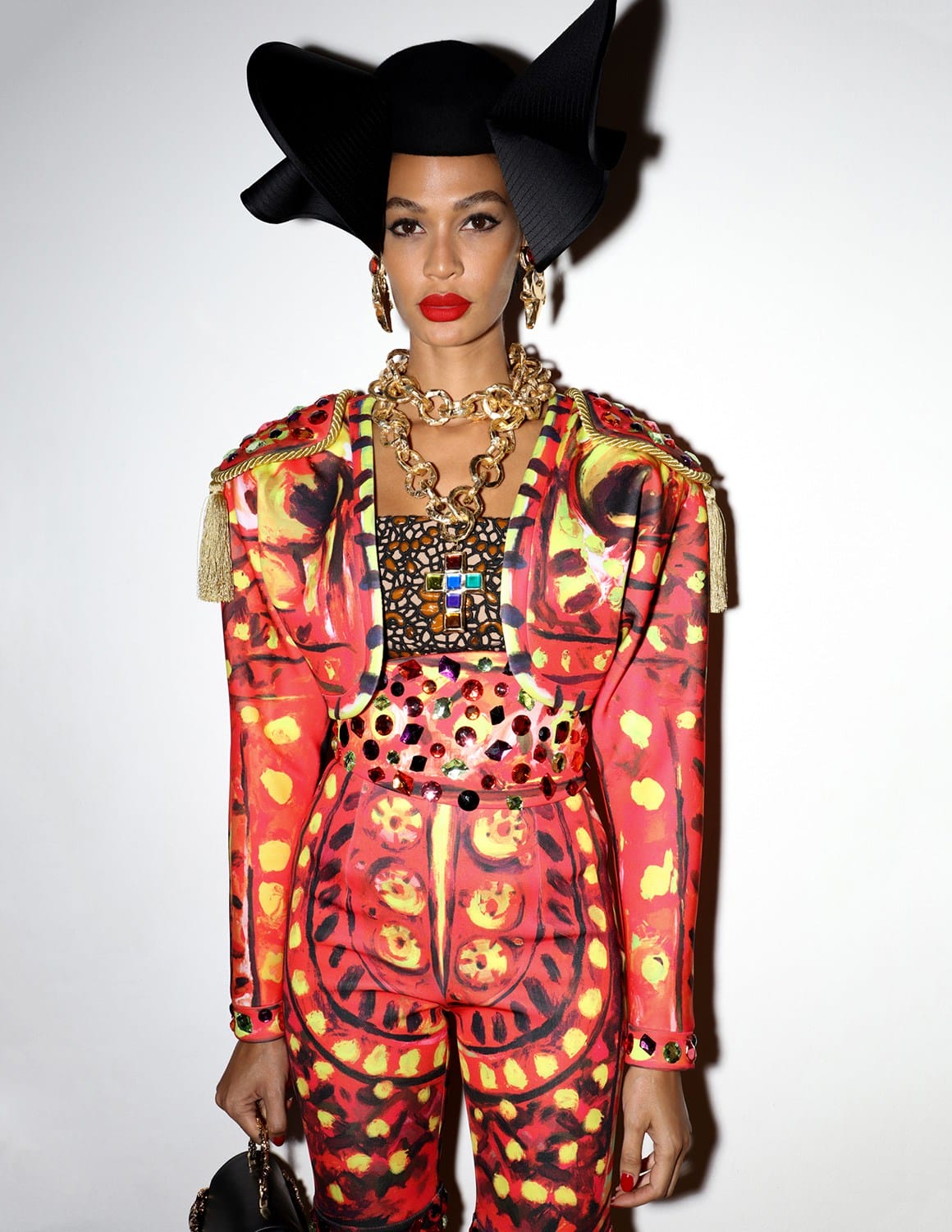 Person wearing a vibrant red and yellow patterned outfit with a large black hat and bold jewelry against a plain background.