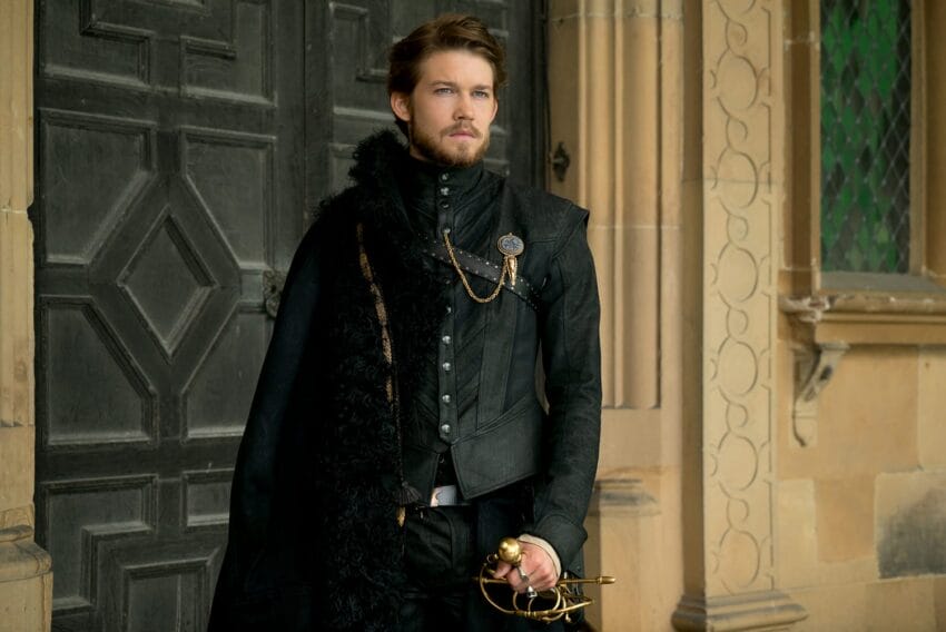 Man in period costume holding a sword, standing in front of a historic building with ornate door and windows.