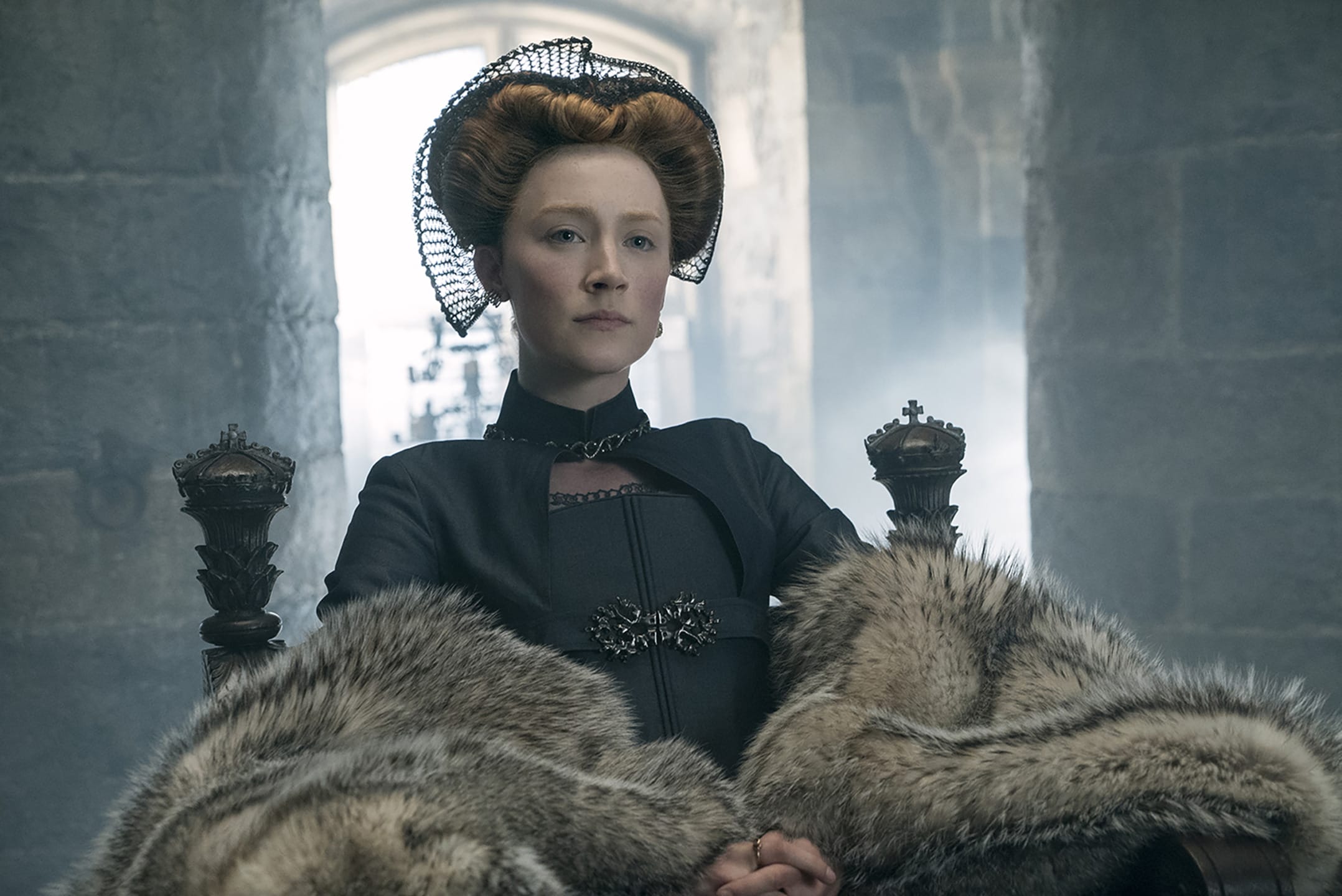 Historical figure in regal attire with fur, sitting on a throne in a dimly lit, stone-walled room.
