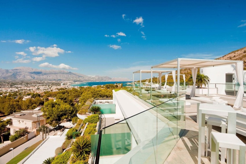 Rooftop terrace with modern furniture overlooking coastal town, hills, and ocean under clear blue sky.