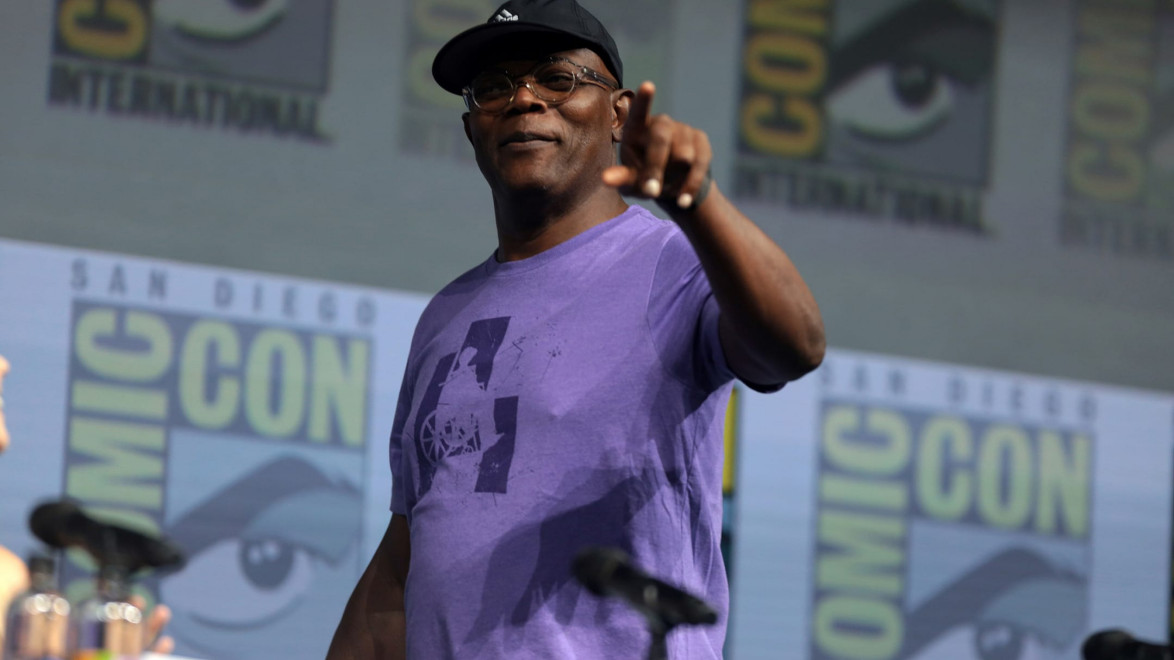 Man in a purple shirt and cap speaking at a Comic-Con event, pointing towards the audience.