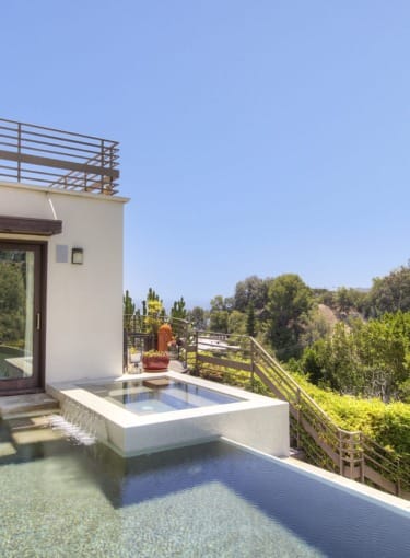 Modern patio with a small pool and hot tub overlooking lush greenery under a clear blue sky.