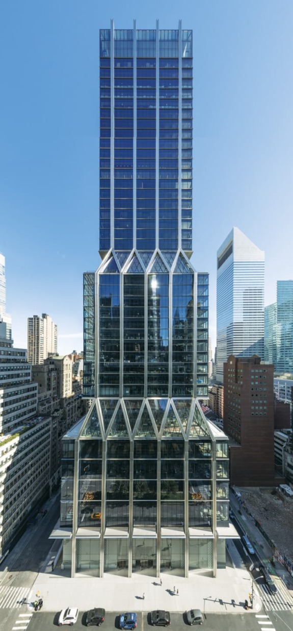 Modern skyscraper with glass facade and geometric design in a cityscape background under a clear blue sky.