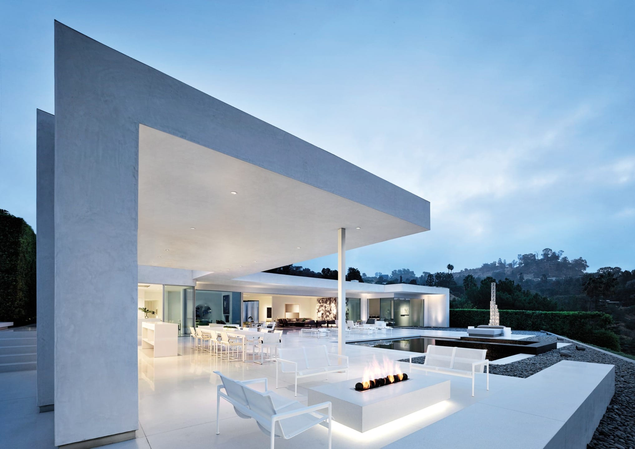 Modern minimalist outdoor patio with white furniture, fire pit, and scenic view of hills at dusk.