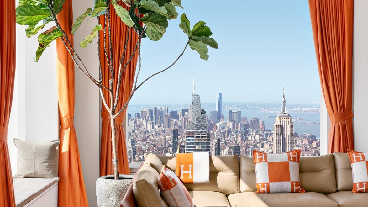 Room with orange curtains and cushions, large plant by window, and cityscape view of skyscrapers in the background.