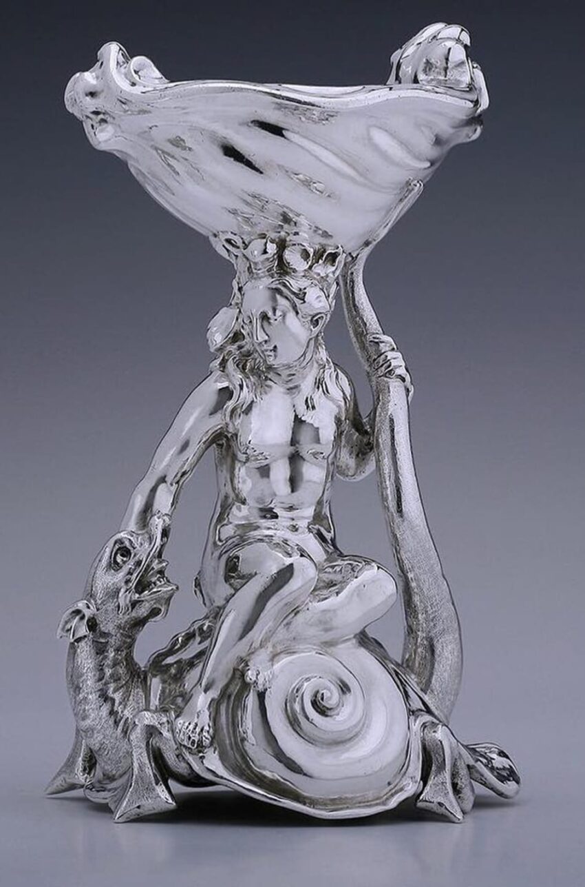 Silver sculpture of a woman sitting on a snail shell, holding a large open clamshell above, with a small creature nearby.
