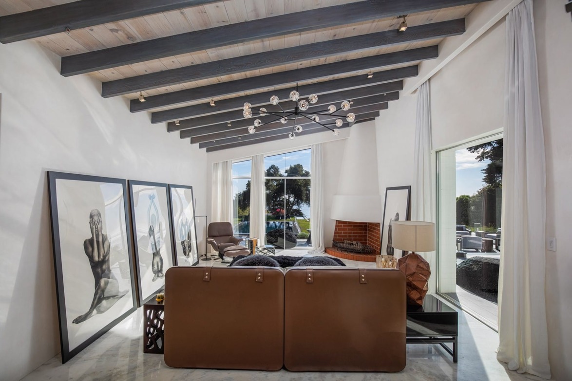 Modern living room with brown sofa, large framed art, exposed beams, and a view of greenery through large windows.