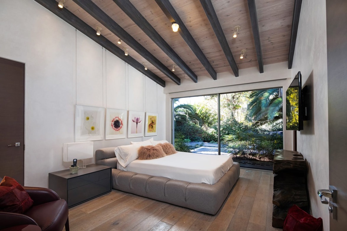 Modern bedroom with wooden ceiling, large window, and nature view; features a bed, nightstands, and wall art.