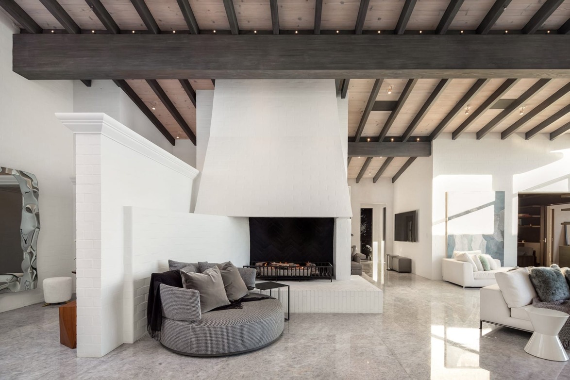 Modern living room with large white fireplace, exposed wooden beams, gray lounge chair, and marble flooring.
