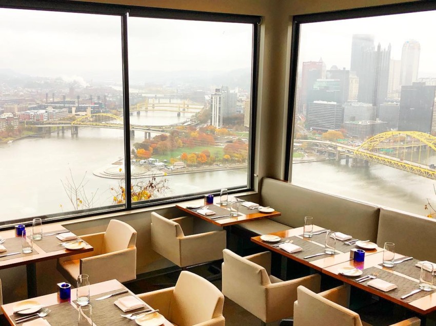 Restaurant with a view of a city skyline and river bridges through large windows, featuring set dining tables.