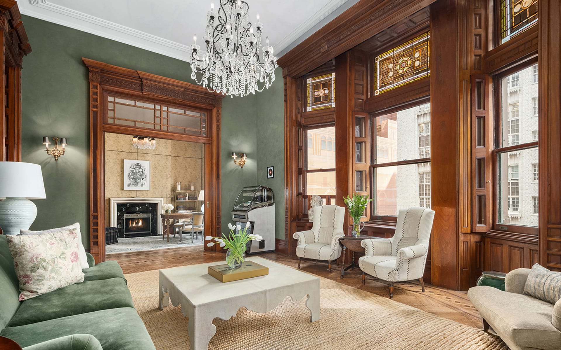 Elegant living room with a chandelier, green walls, large windows, velvet sofa, armchairs, and a view into a dining area with a fireplace.