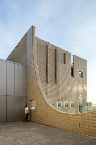 Modern building with unique architectural design and a person standing on a terrace under a clear blue sky.