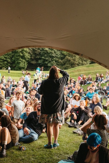 Person speaking to a diverse crowd seated on grassy lawn under a large canopy with trees in the background.