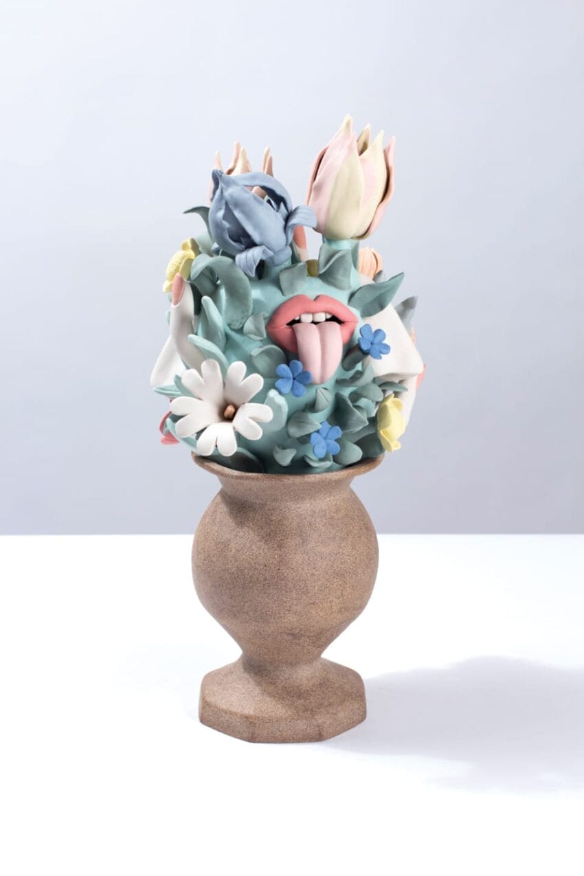 Ceramic vase with whimsical sculpture featuring abstract flowers, lips, and tongue in pastel colors against a light background.