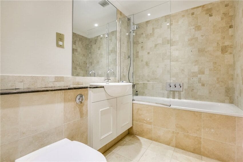 Modern bathroom with beige tiled walls, bathtub with glass screen, white sink vanity, and overhead lighting.