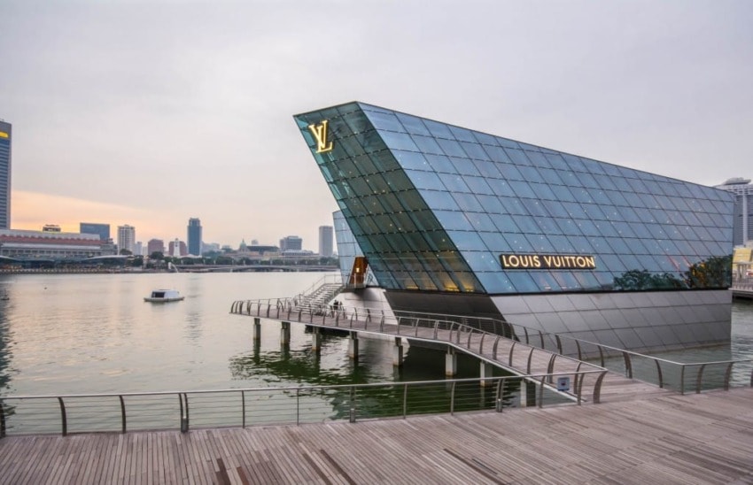 Modern glass building on water with city skyline and boardwalk in the foreground, displaying Louis Vuitton signage.