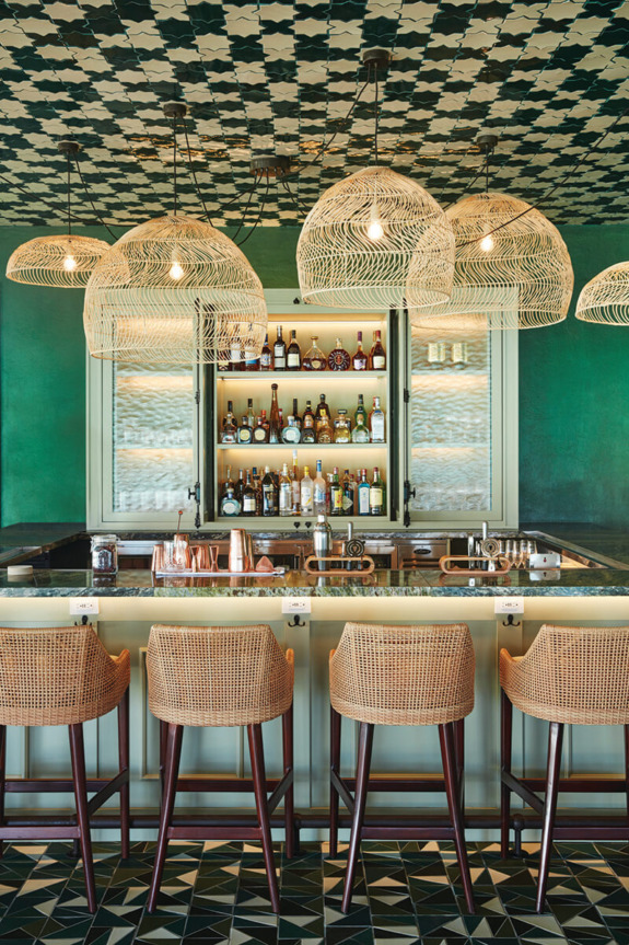 Bar interior with hanging wicker lights, green walls, patterned ceiling, and four wicker bar stools facing a stocked shelf.