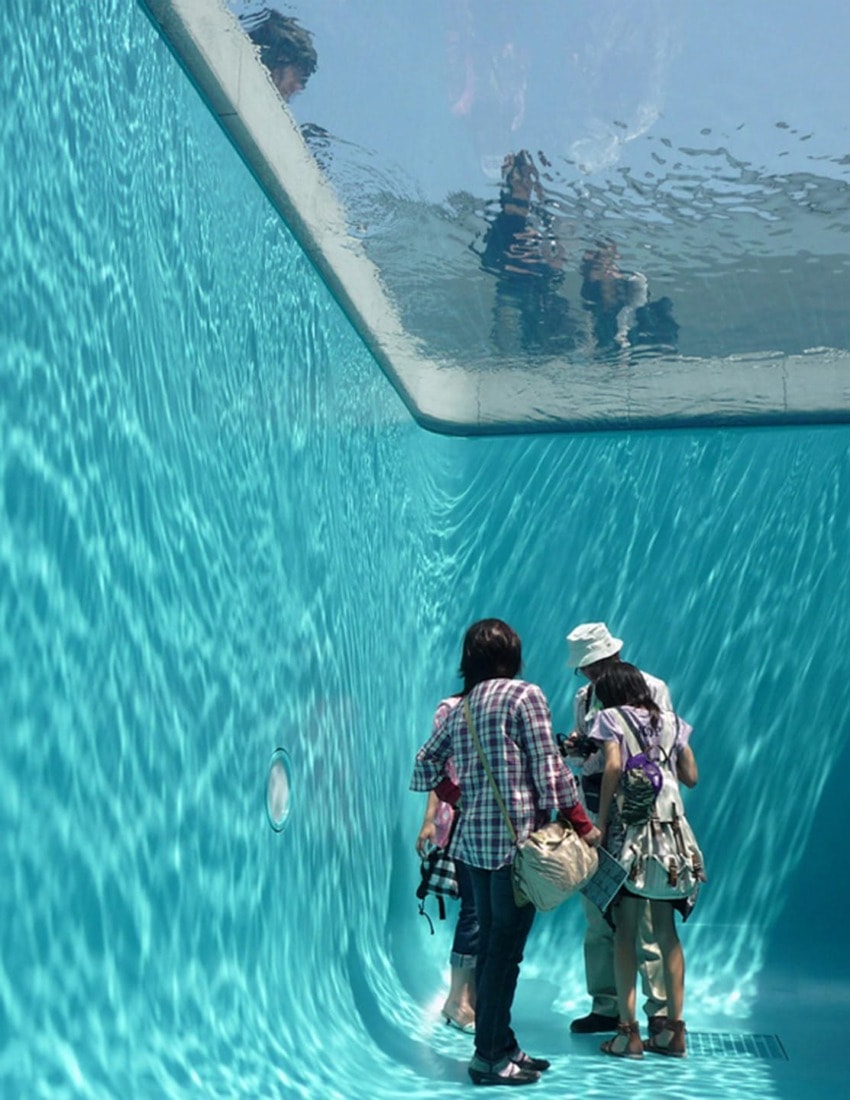 People walking under a transparent pool with reflections on the clear water surface above.
