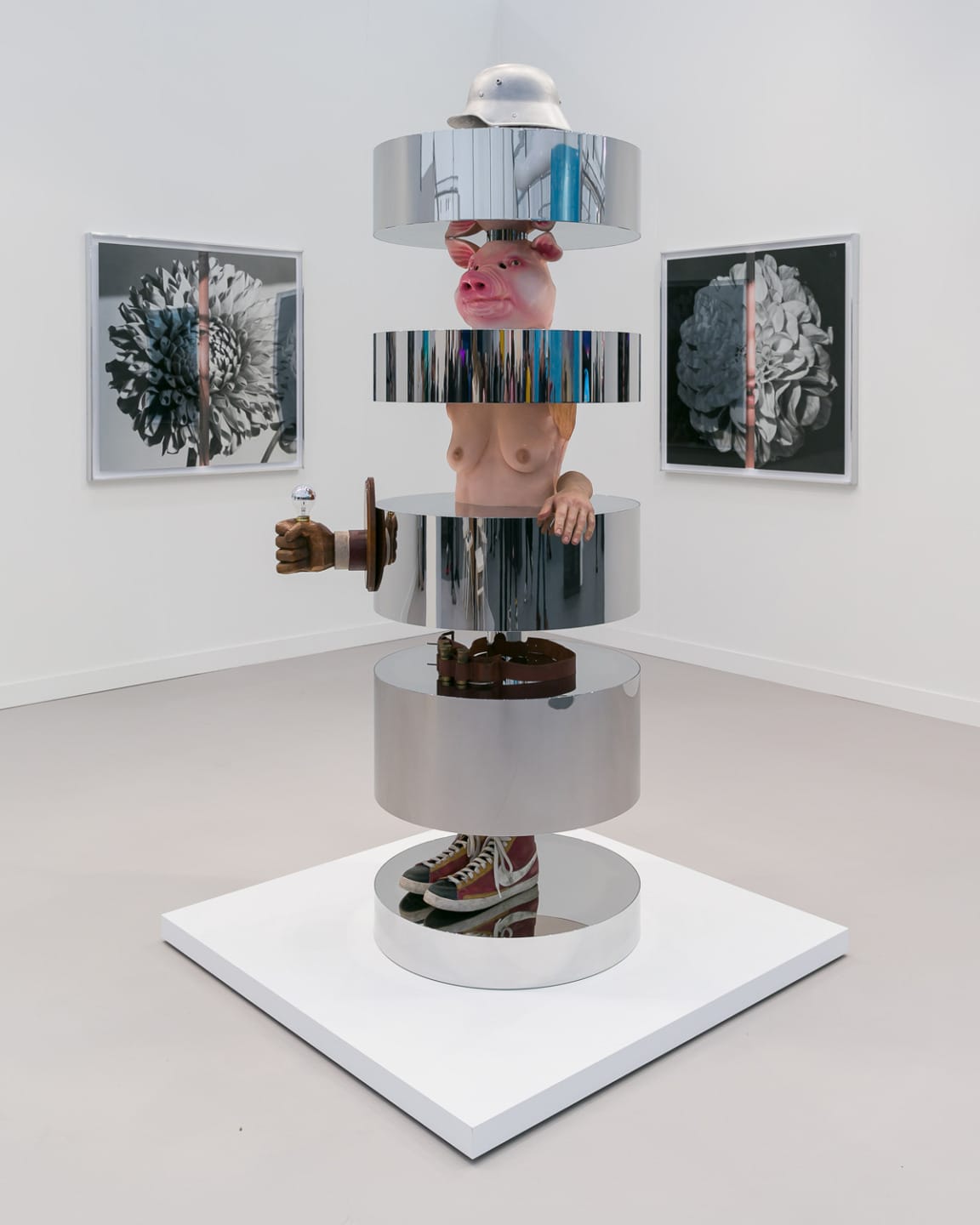 Surreal sculpture with pig head and human arms, segmented metal discs, and sneakers on display in an art gallery.