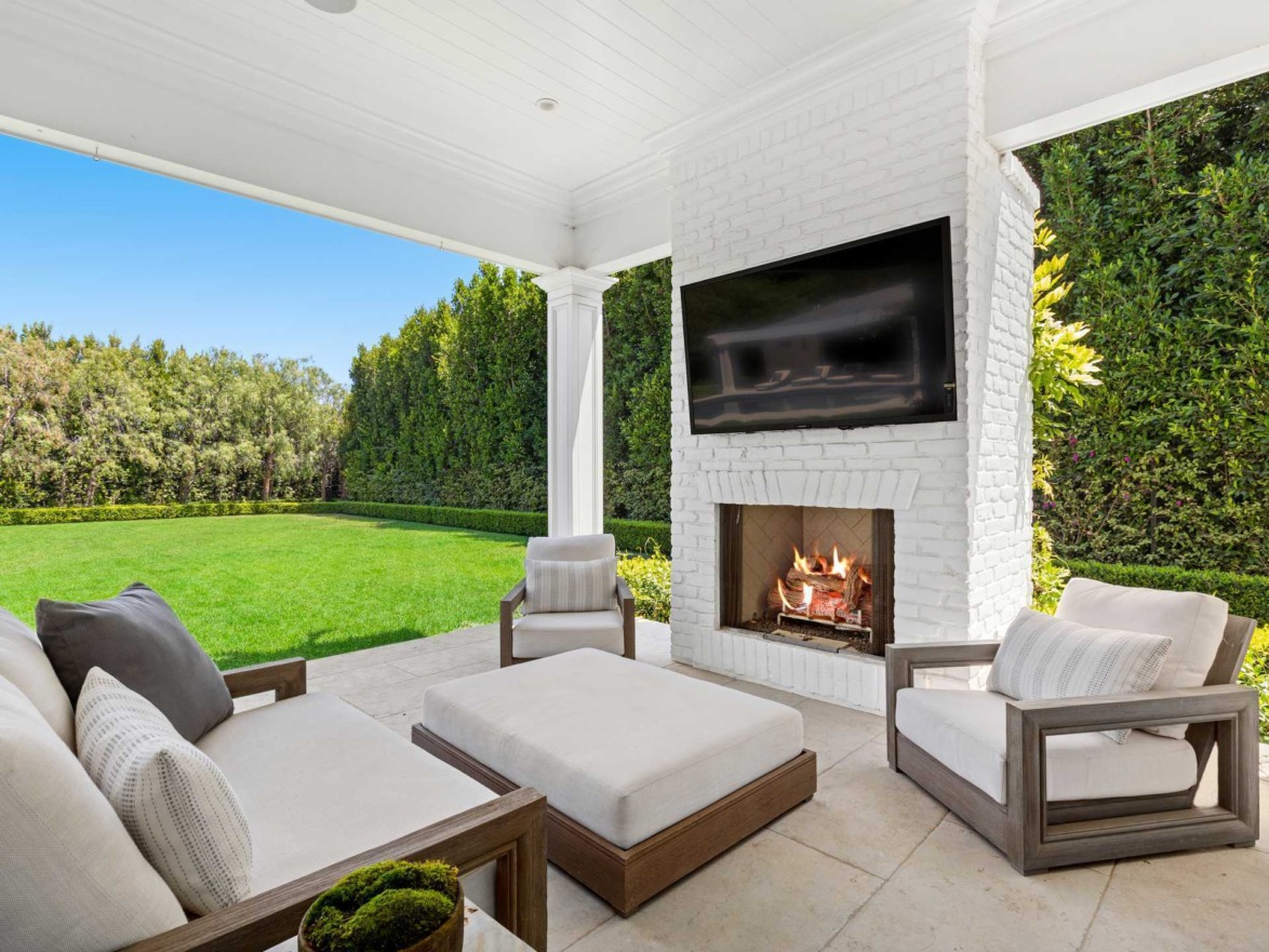 Modern outdoor patio with fireplace, seating area, and large screen TV overlooking a manicured lawn and hedges.