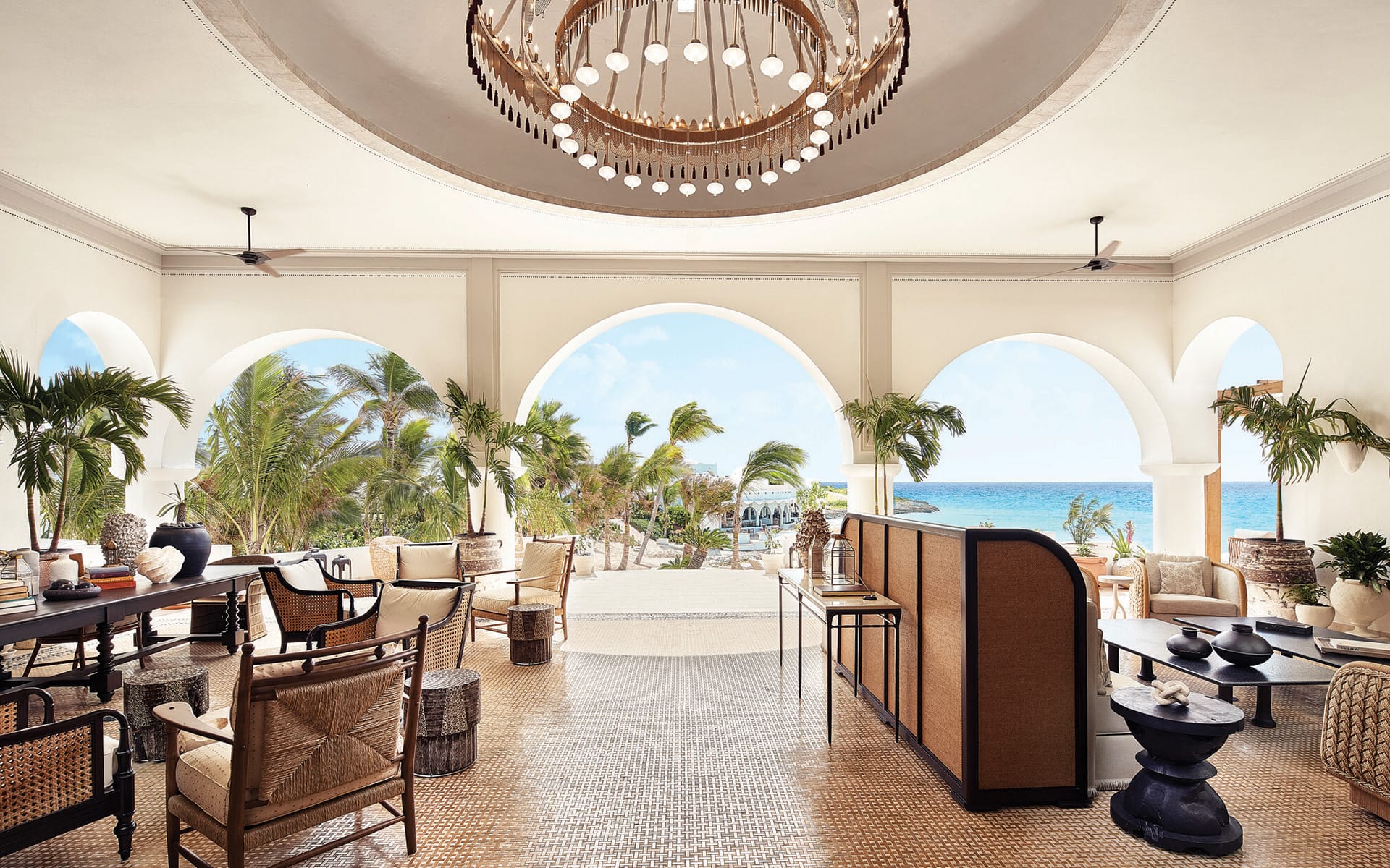 Open-air luxury resort lounge with arched openings, ocean view, wicker furniture, palm trees, and elegant chandelier.