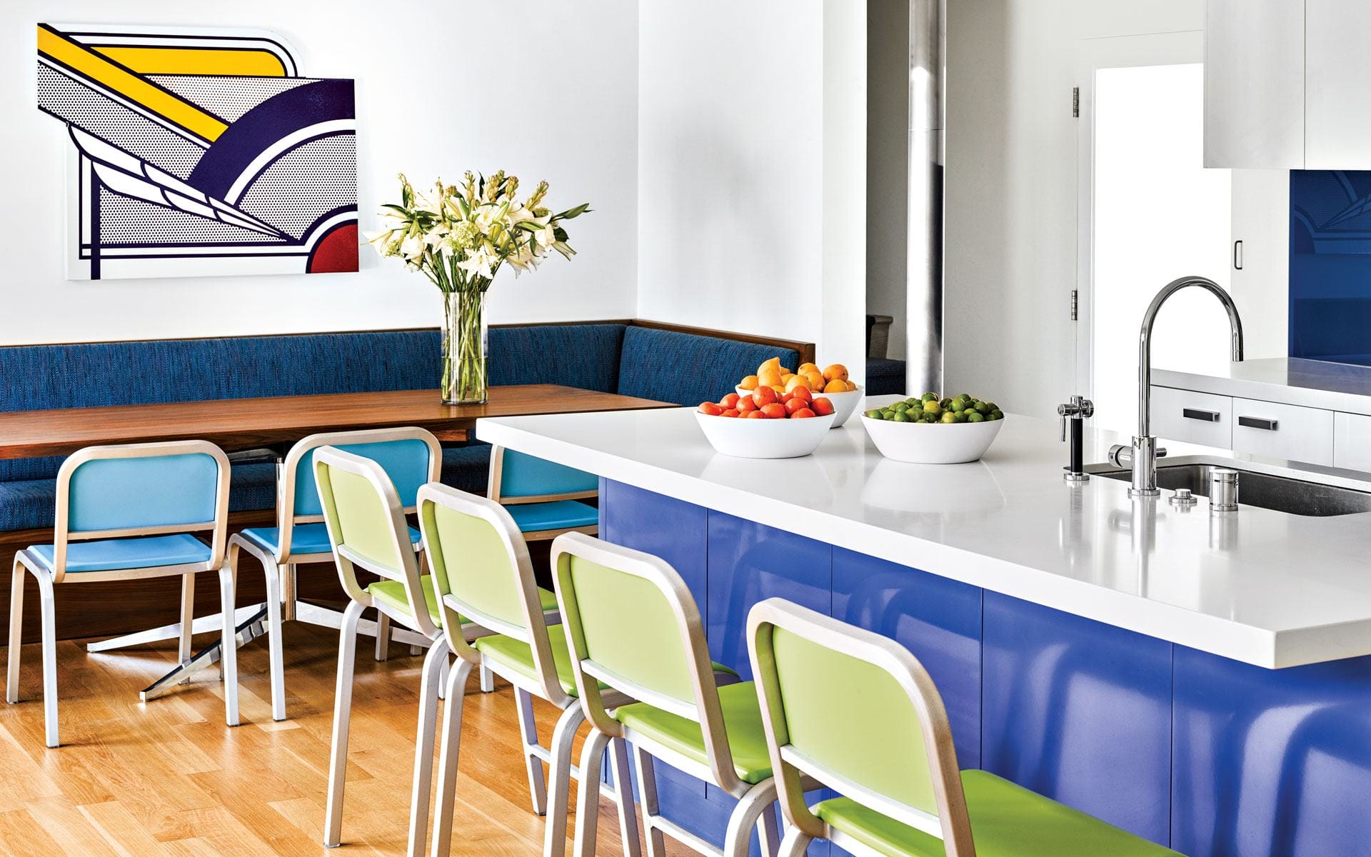 Modern kitchen with blue and green chairs, white countertop, artistic wall decor, and fresh fruit bowls.