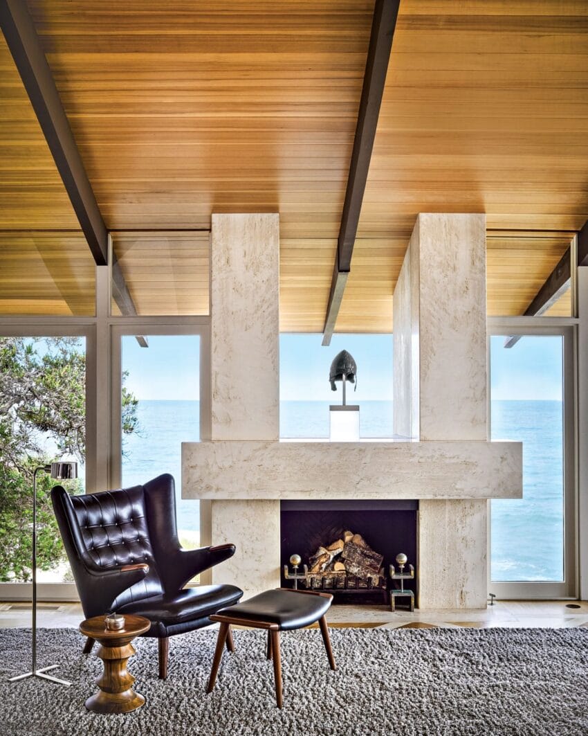 Modern living room with sleek black chair, ottoman, and wooden ceiling, overlooking ocean through large windows beside fireplace.