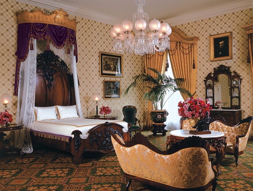 Victorian-style bedroom with ornate wooden furniture, plush gold sofas, chandelier, and floral arrangements.