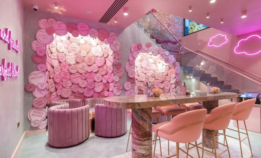 Pink-themed cafe interior with decorative walls, plush seating, marble table, and neon cloud signs. Elegant and modern atmosphere.