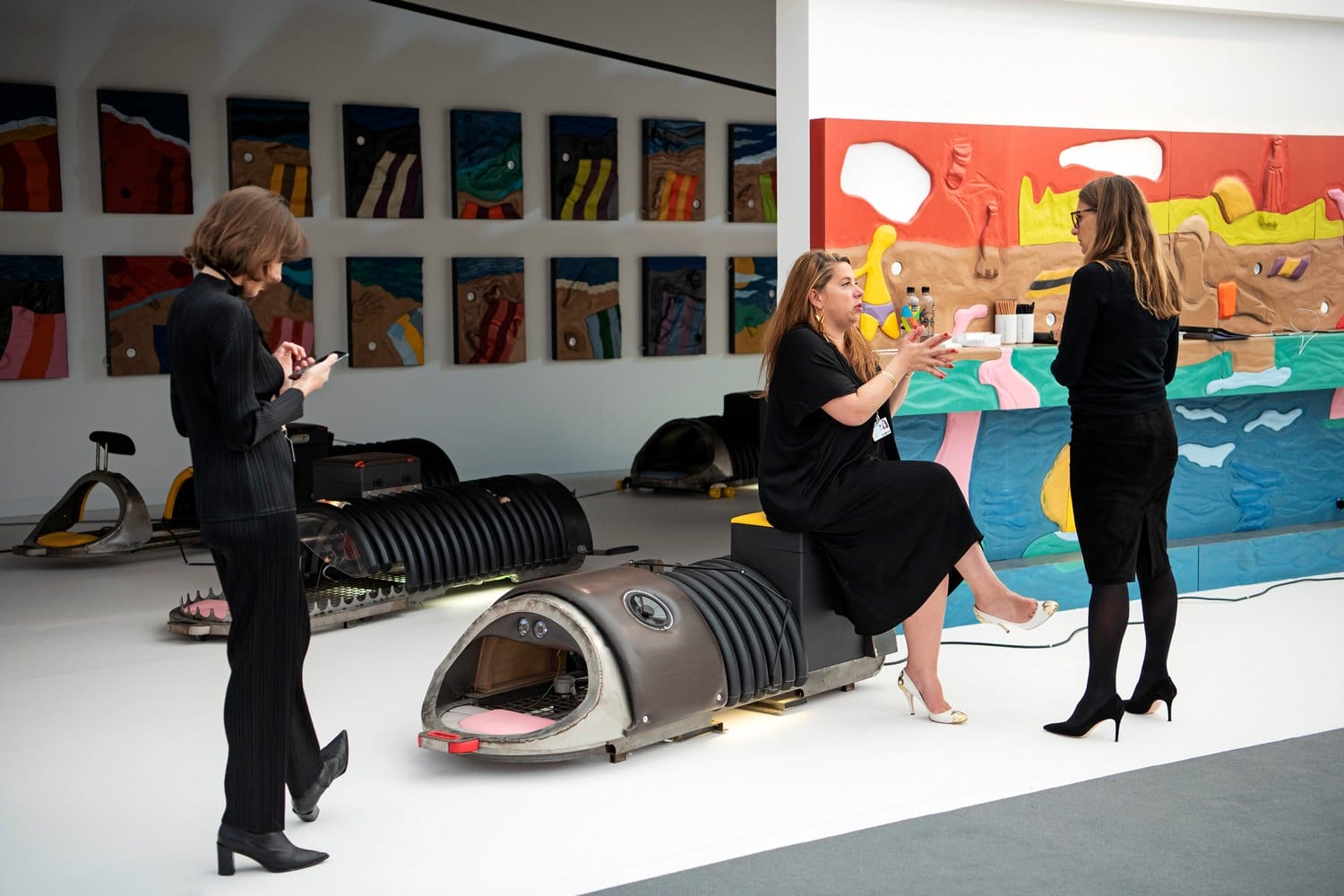 Three women in an art gallery with colorful paintings on walls and futuristic sculptures on the floor.