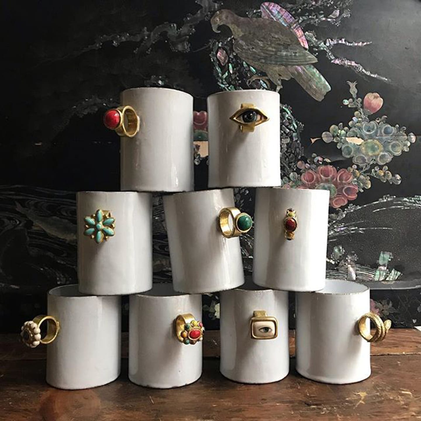 White ceramic mugs with decorative, ring-like handles are stacked in a pyramid against a floral background.