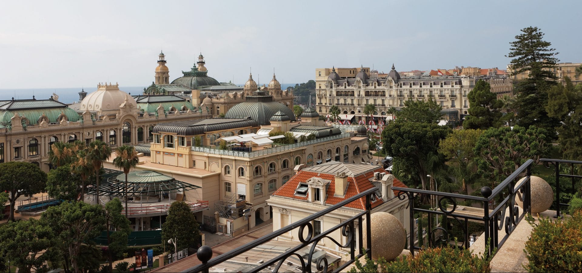 View of historic Monte Carlo skyline with ornate buildings, lush greenery, and a cloudy sky in the background.
