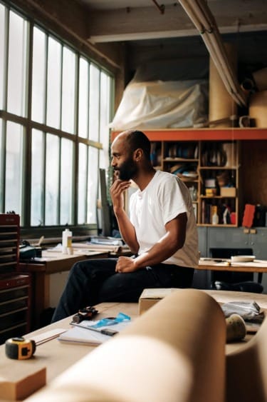 Man sitting on desk in a workshop, looking thoughtful, surrounded by tools and materials, with large windows in the background.