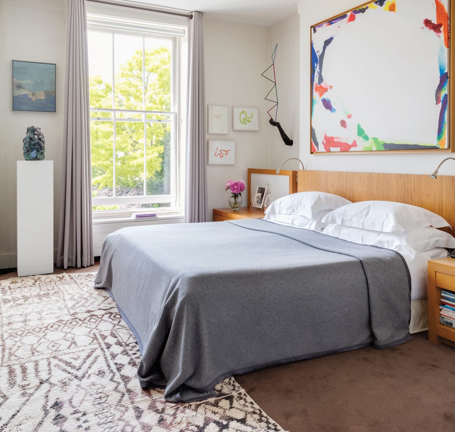 Modern bedroom with large window, gray bedding, abstract art on wall, and decorative rug. Bright and airy atmosphere.