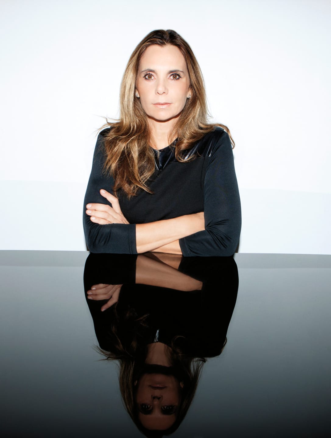 Person with long hair and crossed arms, wearing a black top, sitting at a reflective surface against a white background.