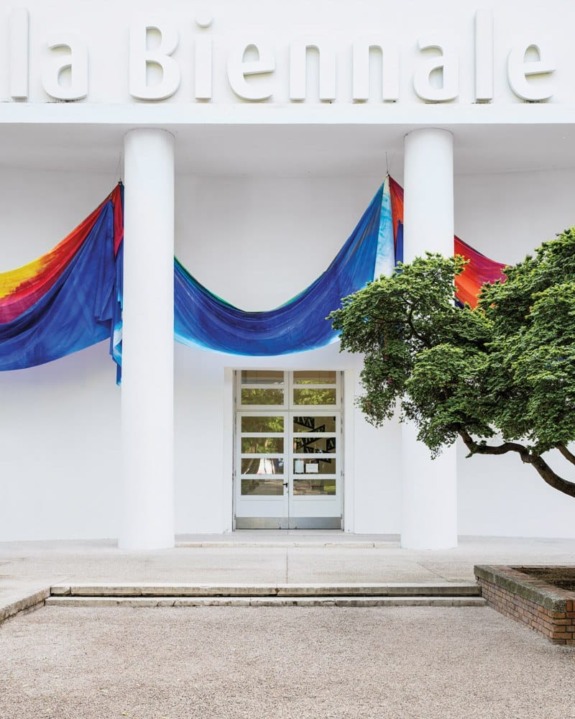 Entrance of a white building with colorful draped fabric and a neatly trimmed tree in the foreground.