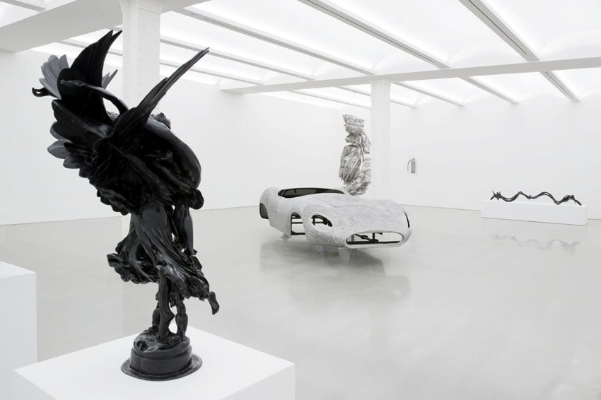 Modern art gallery with sculptures, including a car-inspired piece and abstract black figures on display in a bright space.